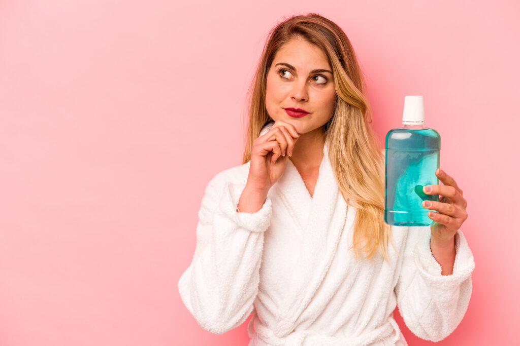 Young caucasian woman holding mouthwash wearing bathrobe isolated on pink background looking sideways with doubtful and skeptical expression.